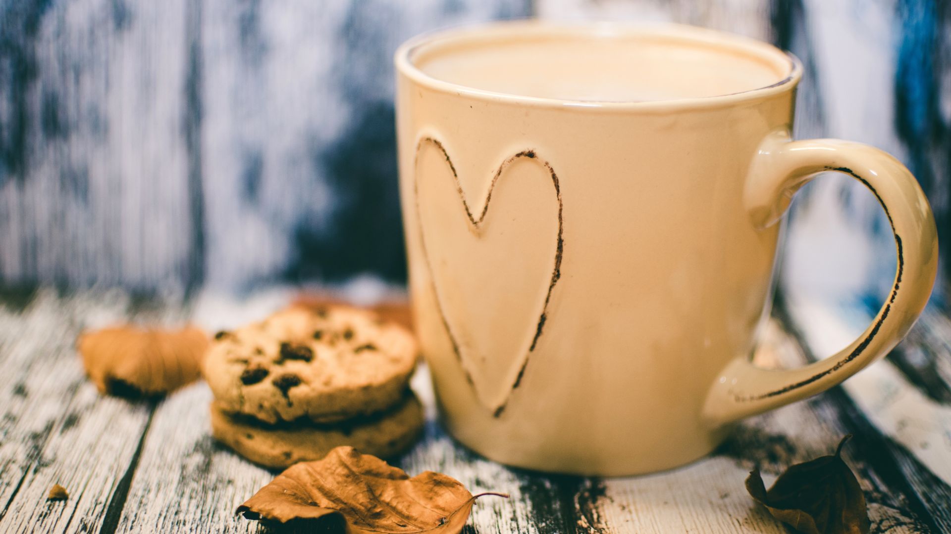 Beige Tasse mit Herzmotiv, umgeben von Keksen und Herbstblättern auf rustikalem Holztisch.