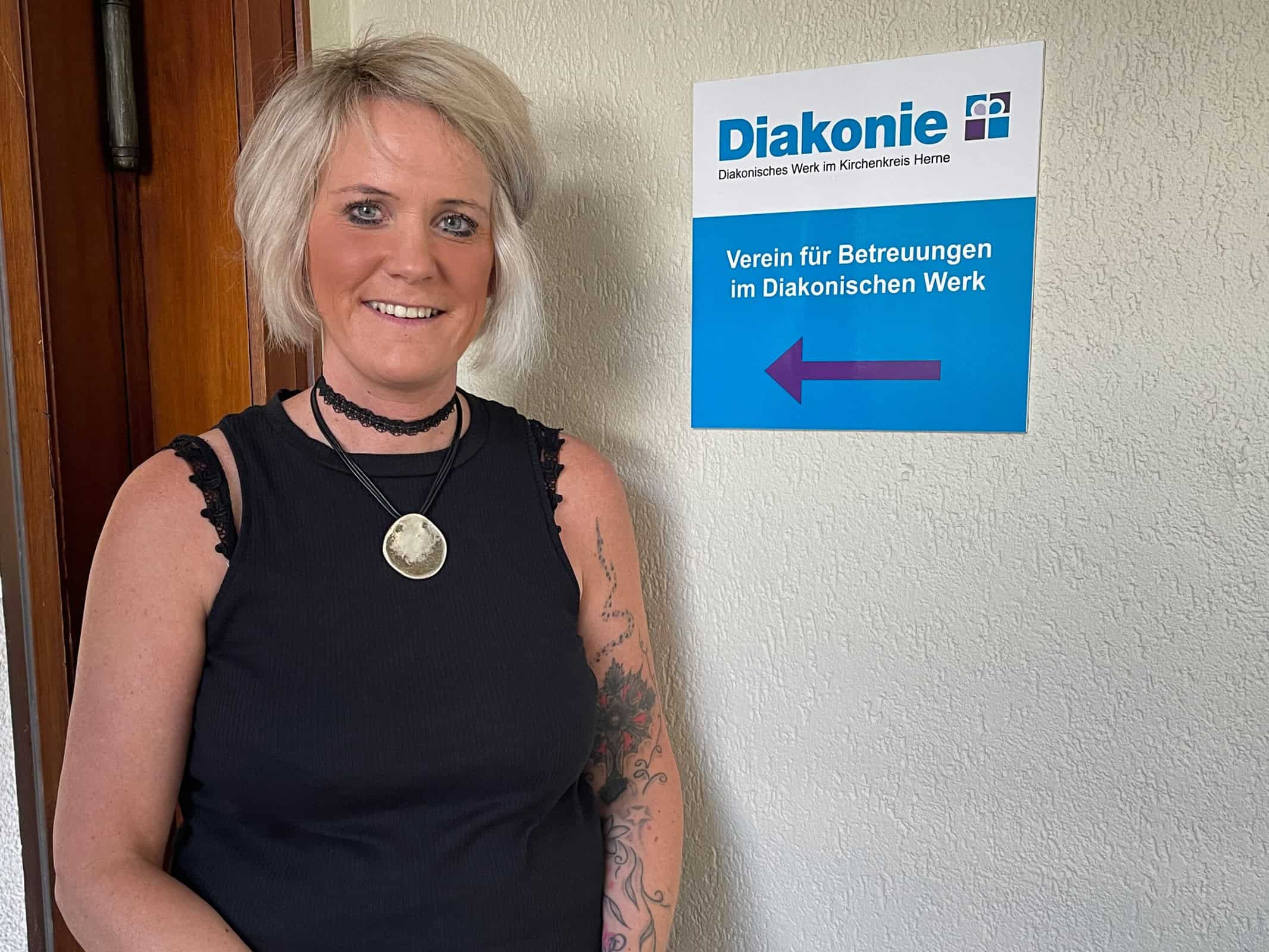 Frau vor Diakonie-Schild in Herne, Deutschland, lächelnd, mit kurzem blondem Haar und schwarzem Oberteil.