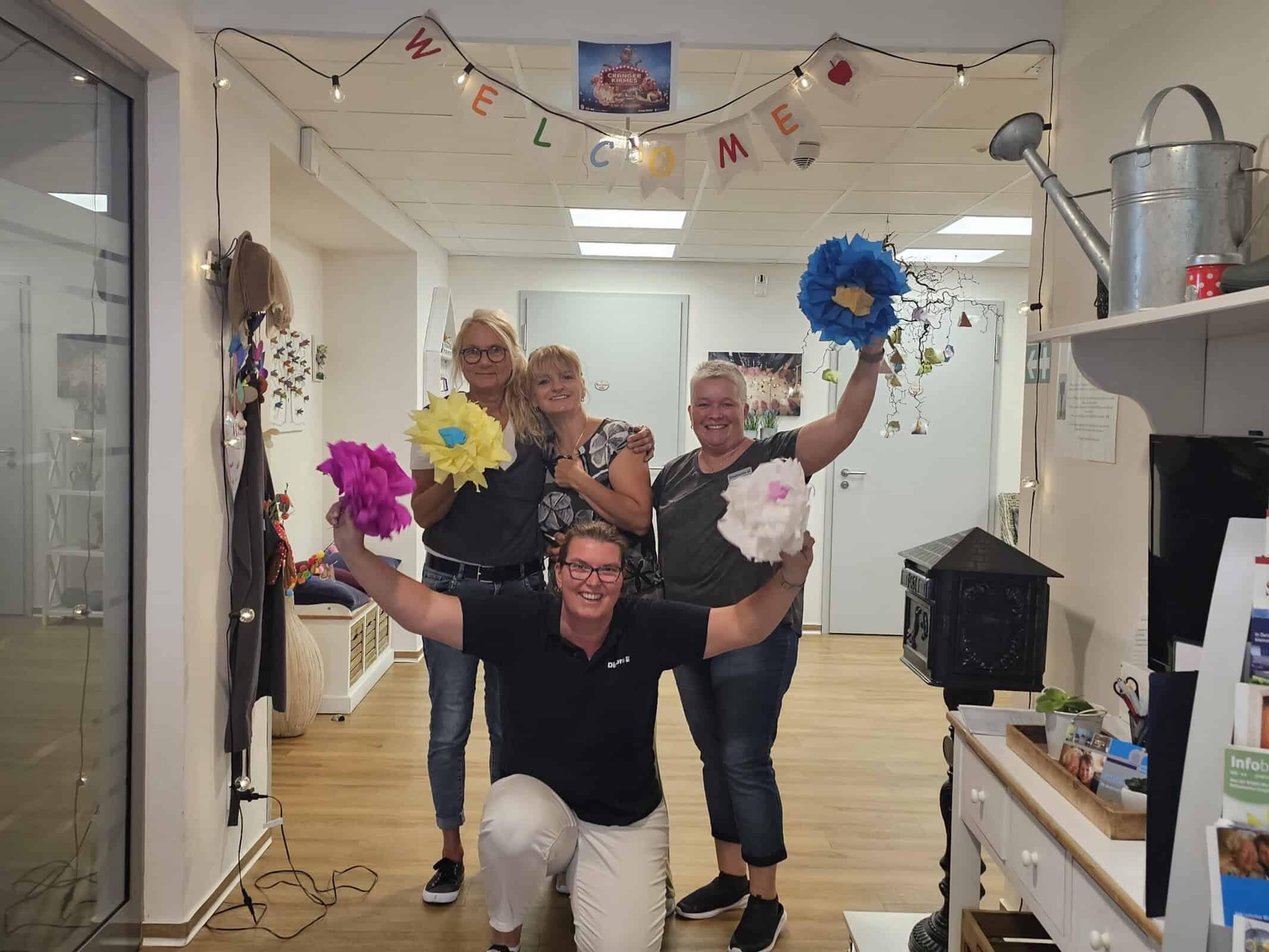 Vier Personen feiern fröhlich mit bunten Papierblumen in einem dekorierten Raum, unter einem Welcome-Banner.