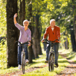 Älteres Paar fährt Fahrrad im Herbstwald, Frau winkt freundlich, sonniger Tag, Fahrradtour, Naturerlebnis.