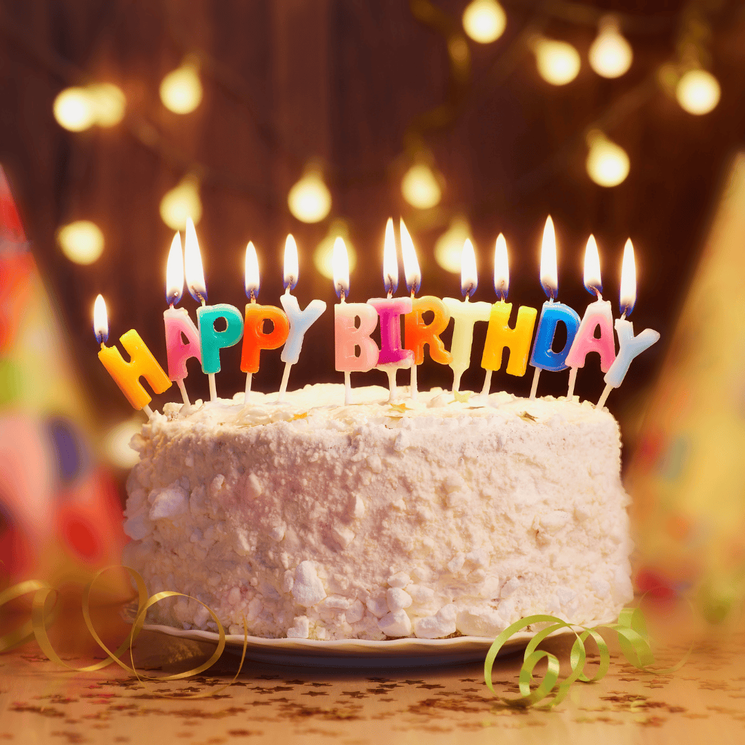 Geburtstagstorte mit brennenden Kerzen, die Happy Birthday buchstabieren, umgeben von Lichtern und Dekorationen.