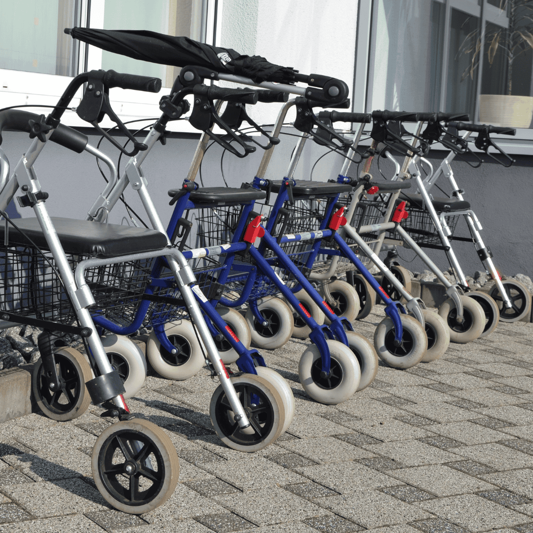 Mehrere Rollatoren nebeneinander auf einem Gehweg vor einem Gebäude geparkt.
