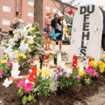 Gedenkstätte mit Kerzen, Blumen und einer Stele mit DU FEHLST in einer städtischen Umgebung.