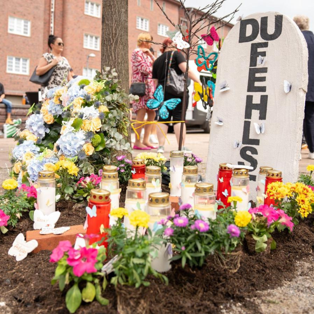 Gedenkstätte mit Kerzen, Blumen und einer Stele mit DU FEHLST in einer städtischen Umgebung.