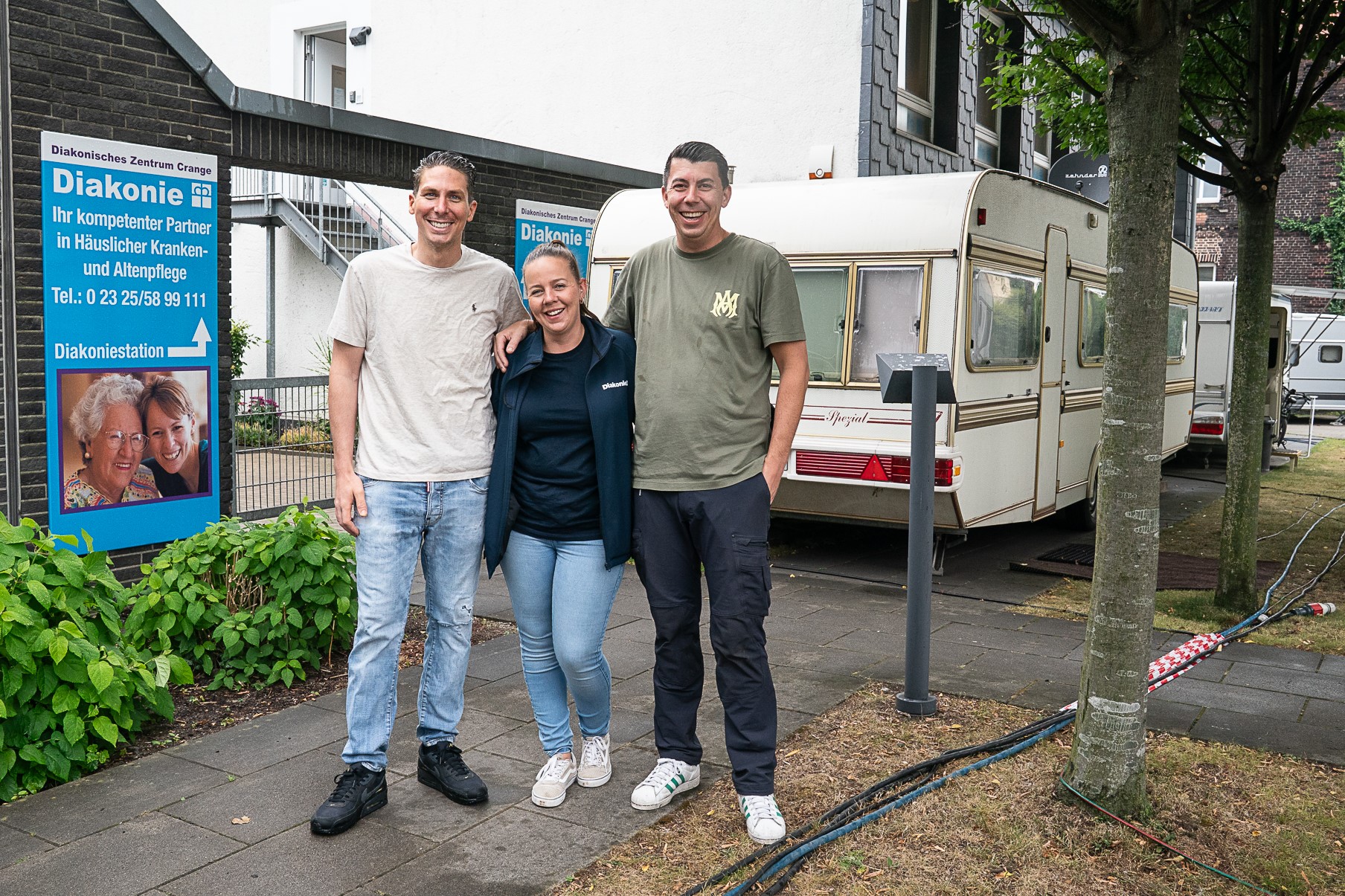 Drei Personen vor einem Schild der Diakonie Crange, neben einem Wohnwagen in einer städtischen Umgebung.