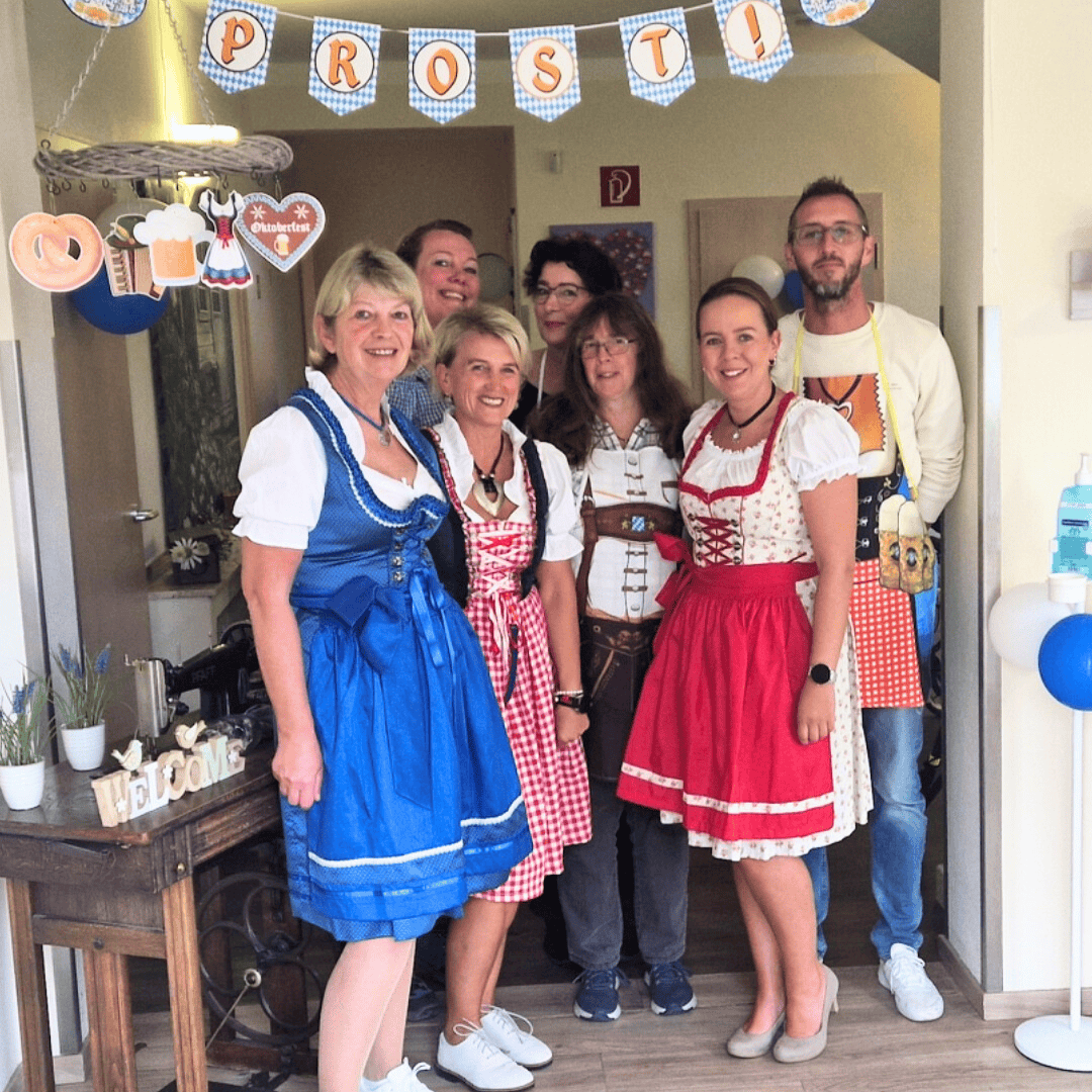 Personengruppe in traditioneller bayerischer Tracht bei Oktoberfest-Feier mit Prosit-Girlande im Hintergrund.