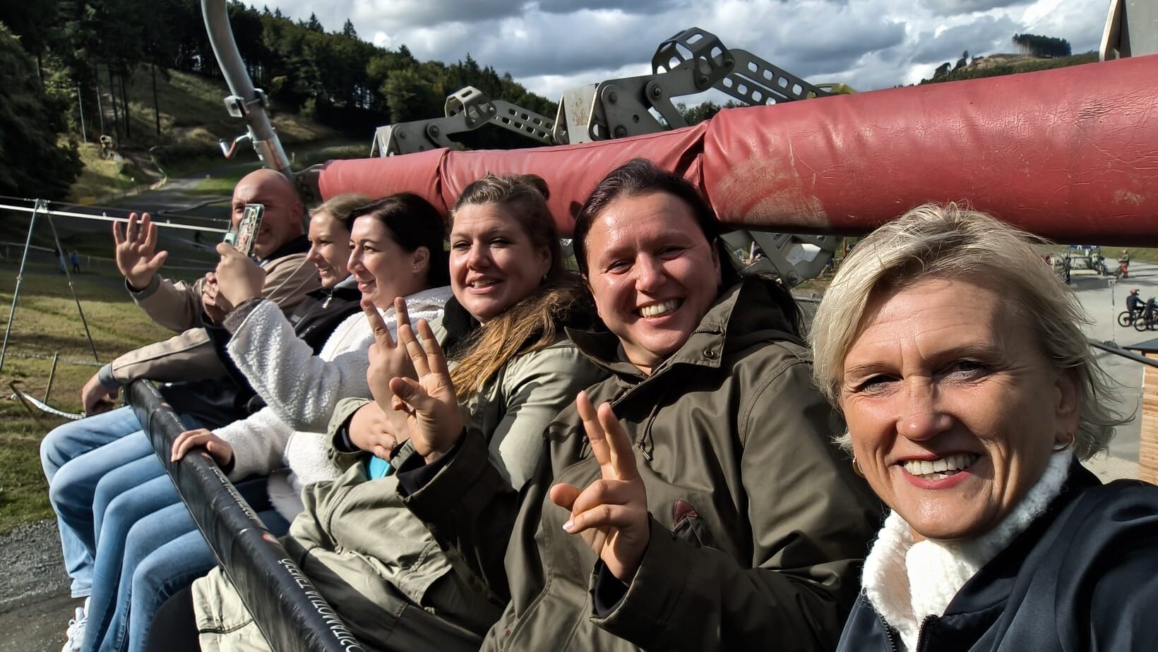 Gruppe von Menschen auf einem Sessellift lächelt in die Kamera, sonniger Tag mit bewölktem Himmel im Hintergrund.