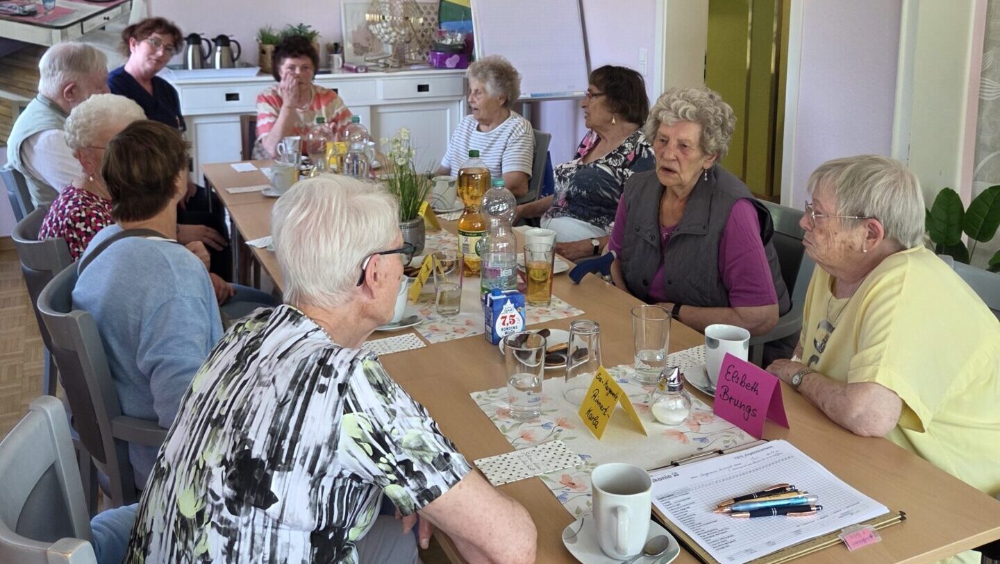 Ältere Menschen sitzen um einen Tisch in einem Gemeinschaftsraum und unterhalten sich in freundlicher Atmosphäre.