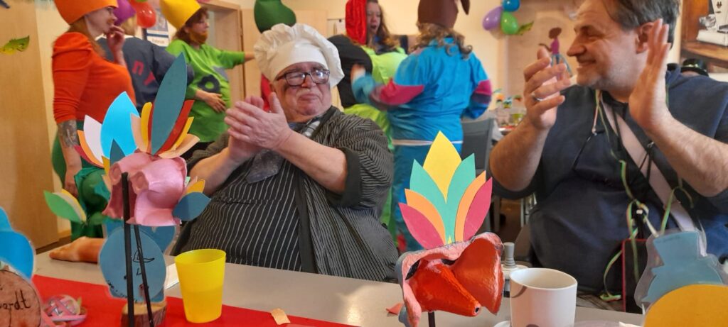 Älterer Koch klatscht mit Männern bei bunter Party in farbenfrohen Kostümen und Dekorationen im Hintergrund.