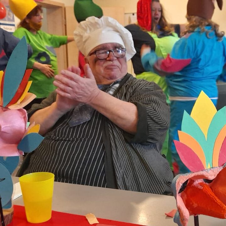 Älterer Koch klatscht mit Männern bei bunter Party in farbenfrohen Kostümen und Dekorationen im Hintergrund.