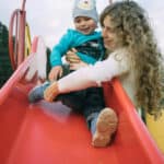 Mutter spielt mit Kind auf roter Rutsche im Park, beide lächeln glücklich. Outdoor-Spaß auf dem Spielplatz.