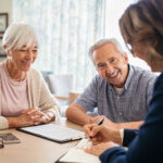 Älteres Paar lächelt bei Beratungsgespräch, während Beraterin Notizen macht.