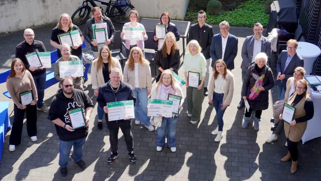 Gruppe von Menschen hält Urkunden bei einer Outdoor-Veranstaltung in Deutschland, sonniger Tag, feierliche Stimmung.