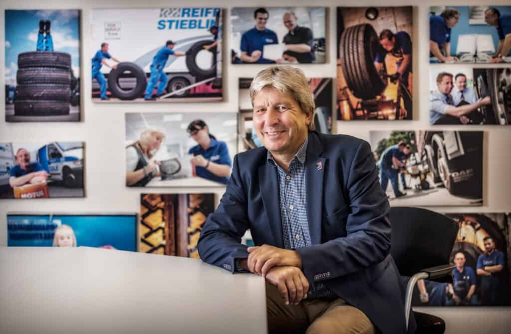Mann in Anzug lächelt vor Wand mit Fotos von Reifenwechsel und Werkstätten.